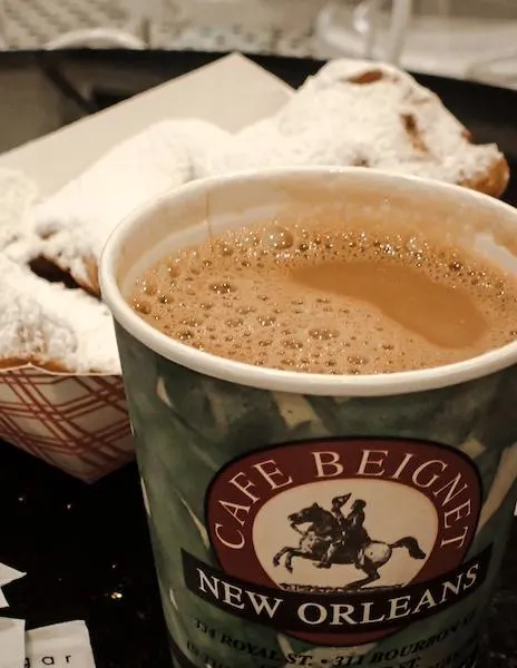 A cup of cafe au lait from Cafe Beignet in New Orleans, prominently displayed with the café's logo, alongside a tray of beignets heavily dusted with powdered sugar in the background.