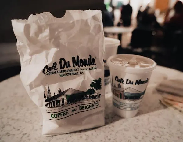 A branded bag of Cafe Du Monde coffee and beignets next to two cups of coffee with the cafe’s logo, set on a table in a dimly lit setting.
