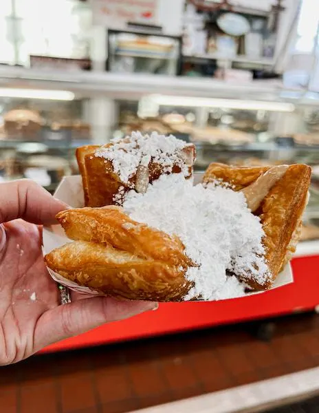 a hand holding a tray of golden beignets heavily topped with powdered sugar, showcased in front of a cafe counter.