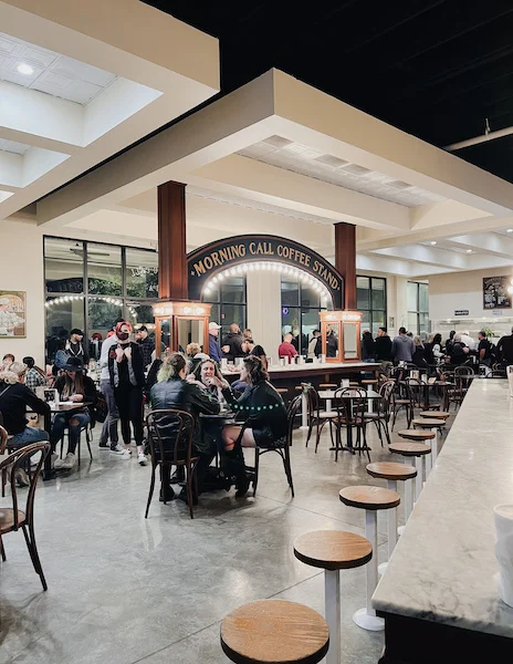 Busy interior of Morning Call Coffee Stand, featuring customers seated at tables and a long marble bar, under a warmly lit, spacious setting.