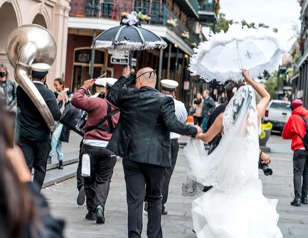 Bride and groom leading a Second Line parade through the French Quarter in New Orleans, dancing with decorated umbrellas as a brass band plays.