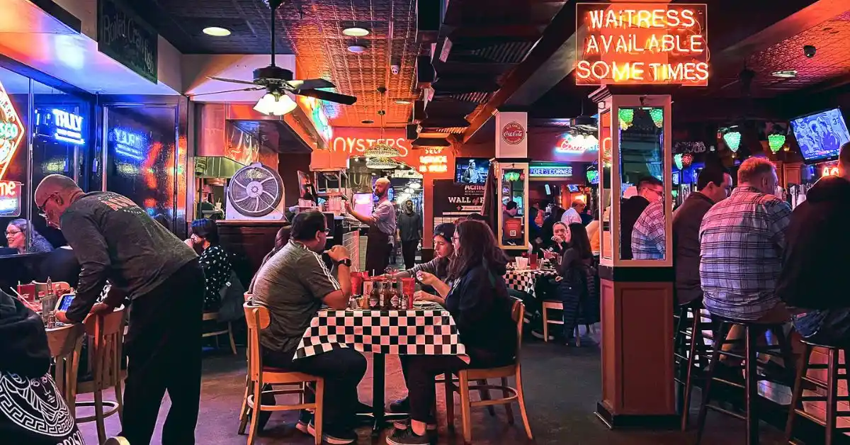 featured image: Casual restaurant scene in New Orleans with neon signs, checkered tablecloths, and diners enjoying local food. The warm glow and lively atmosphere capture classic places to eat in New Orleans.