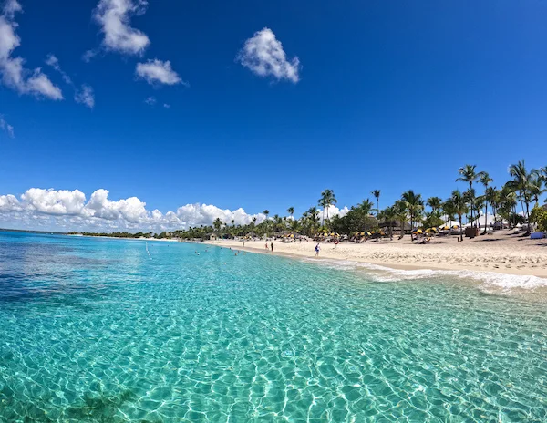A stunning beach on Isla Catalina, Dominican Republic, featuring clear turquoise water, white sand, and people relaxing under palm trees and umbrellas against a bright blue sky.