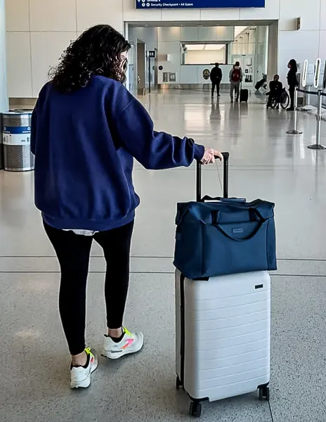 Kathy walking through airport with carry-on suitcase and personal item bag stacked on top before security checkpoint.