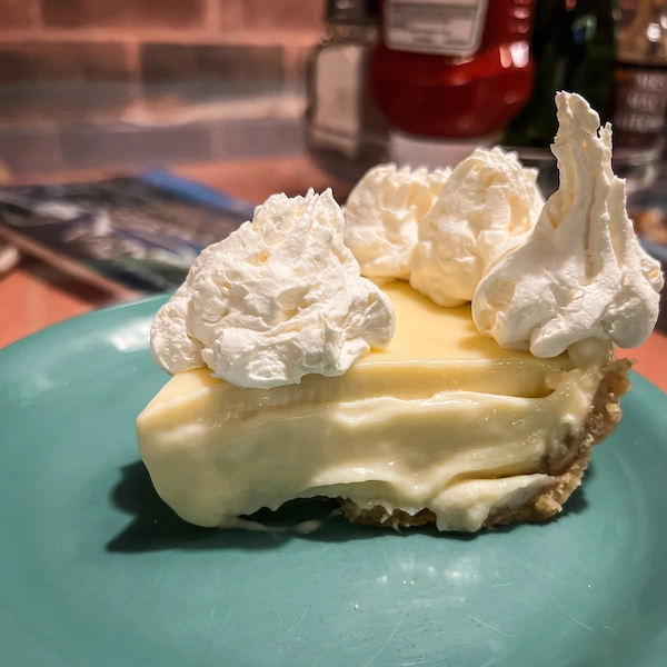 A slice of key lime pie from Mrs. Mac's Kitchen in Key Largo, featuring a creamy filling and topped with a generous dollop of whipped cream, on a teal plate.