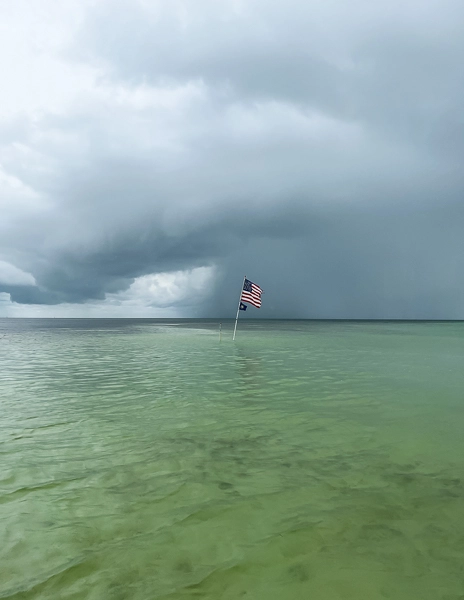 An American flag standing in shallow turquoise waters at a sandbar in the Florida Keys, with a dramatic stormy sky in the background.