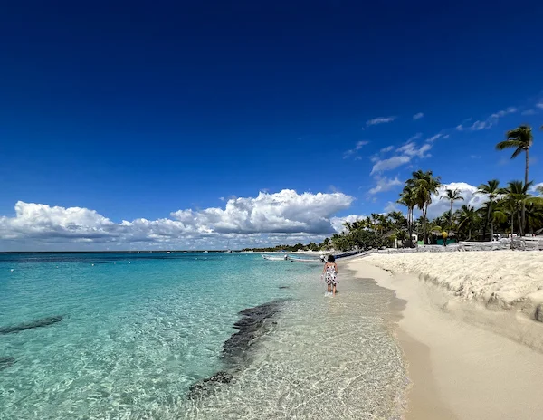 A serene beach on Isla Catalina, Dominican Republic, with clear turquoise water, white sand, and kathy (woman) walking along the shore under a bright blue sky dotted with clouds and palm trees