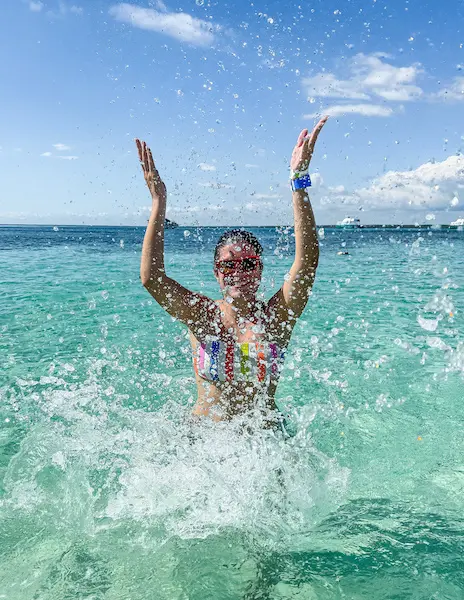Kathy (a woman) splashing in the clear turquoise waters of Isla Catalina, Dominican Republic, with her arms raised joyfully, wearing sunglasses and a colorful bikini, under a bright blue sky with a few clouds.