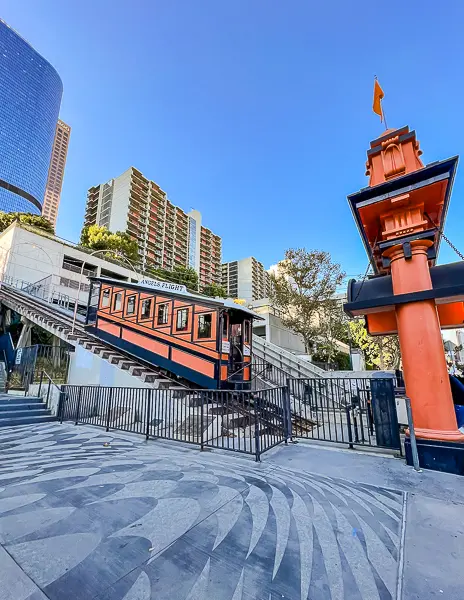 Street-level view of Angels Flight Railway in Downtown LA, showing the orange railcar, entrance gate, and surrounding city buildings.
