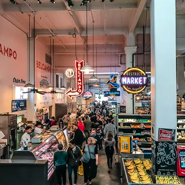 Crowds walking through Grand Central Market in Downtown LA, with neon vendor signs and fresh produce stands lining the walkway.