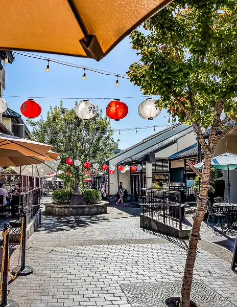 Hanging red and white paper lanterns in a sunny courtyard at Japanese Village Plaza in Little Tokyo, surrounded by shops, trees, and outdoor seating.