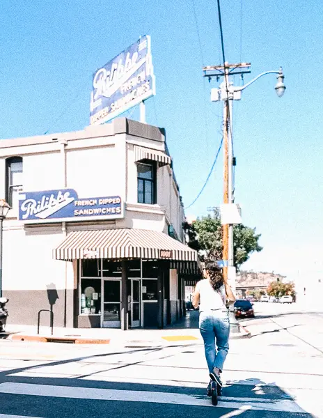 A person (kathy) riding a scooter past Philippe’s French Dip Sandwiches in Downtown Los Angeles, with the restaurant’s vintage neon sign visible above.