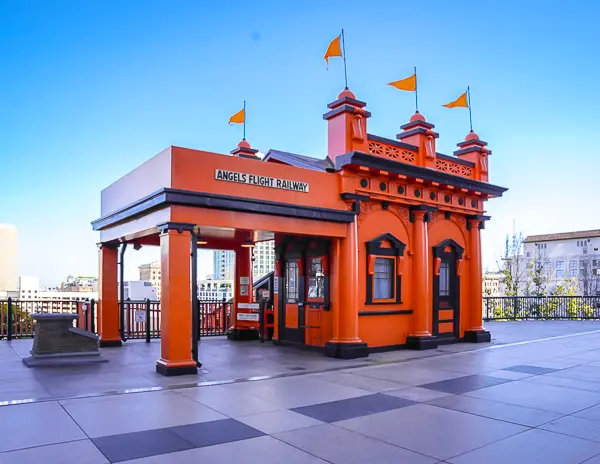 The bright orange upper station of Angels Flight Railway in Downtown LA, with flags flying above and a clear view of the city skyline behind it.