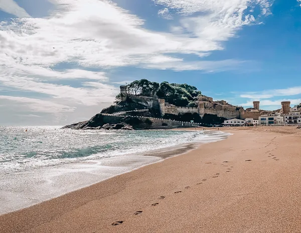 The shore to the left and sand to the right with foot prints leading up to castle ruines perched on a hill in Tossa del Mar