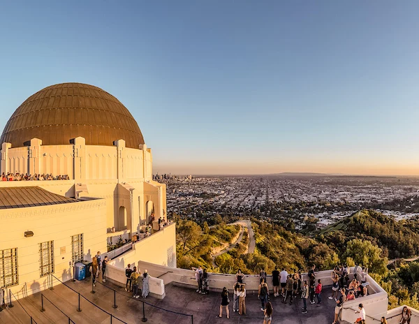 a breathtaking view from Griffith Observatory at sunset. The foreground shows visitors gathered on the observatory's viewing terraces, enjoying the expansive cityscape of Los Angeles stretching into the horizon. The observatory's iconic white dome and elegant architecture contrast beautifully with the golden hues of the setting sun