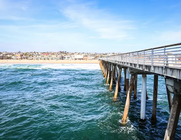 the Hermosa Beach Pier, extending gracefully into the vibrant blue Pacific Ocean. The pier, supported by robust wooden pilings, leads the eye toward the bustling Hermosa Beach shoreline, where a lively beach community is visible.