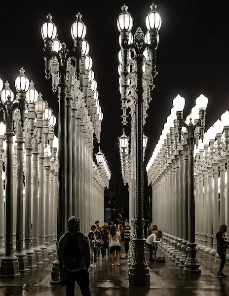 A night view of the Urban Lights at LACMA, beautifully illuminated, with visitors walking among the densely arranged historic street lamps that cast a dramatic glow.