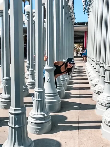 A playful moment captured at the Urban Lights display at LACMA, with a woman (my bff) peeking through the rows of white street lamps, highlighting the interactive nature of this popular art installation.