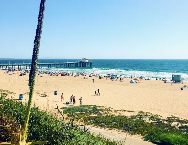  picturesque view of Manhattan Beach in California. The scene features a wide, sandy beach bustling with people enjoying a sunny day. A distinctive pier, extending into the ocean