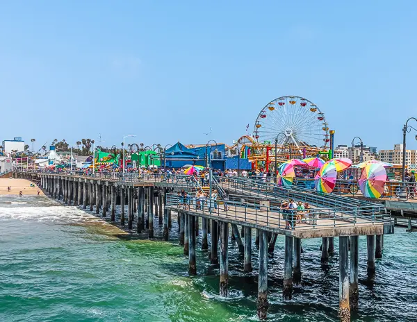 This image captures the vibrant and colorful Santa Monica Pier in California. The pier is bustling with activity, featuring a Ferris wheel, roller coaster, and various amusement park attractions. Large colorful umbrellas and a crowd of visitors add to the festive atmosphere. The ocean in the background and clear blue skies make it a quintessential Southern California scene.