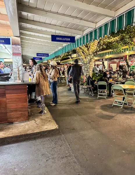 a busy walkway inside the Farmers Market, lined with various stalls selling shellfish and other goods. People are browsing and interacting with vendors, creating a lively marketplace atmosphere.