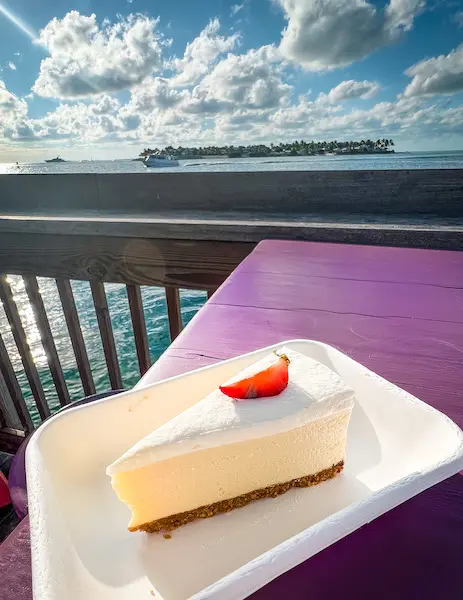 A slice of key lime pie with a smooth topping at Sunset Pier, served on a paper plate with a scenic ocean backdrop.