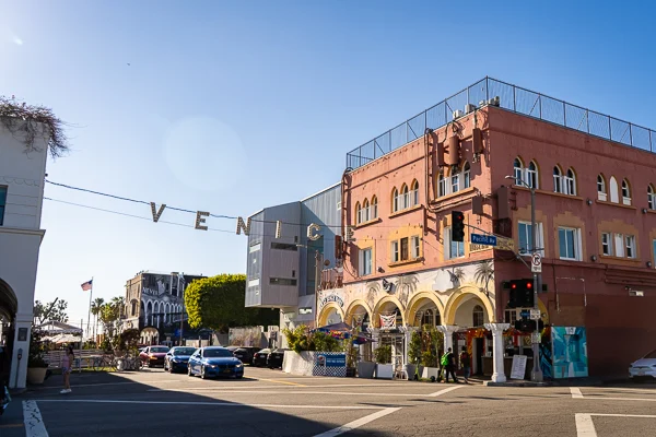 This image shows a vibrant street scene at Venice Beach, California, featuring the iconic "VENICE" sign hanging across the street.