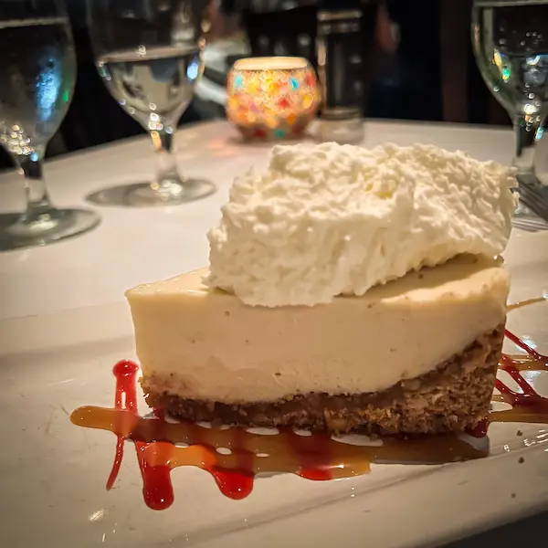 A slice of key lime pie from Ziggy and Mad Dog's topped with whipped cream, served on a white plate with drizzled caramel and raspbery sauce with a decorative candle in the background on the table.