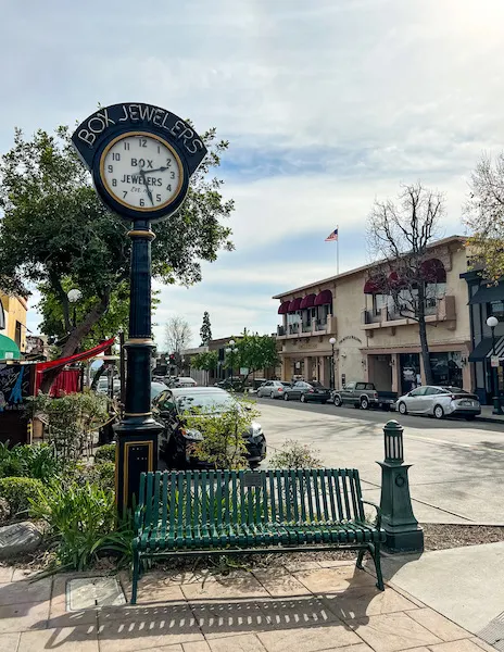 Historic Box Jewelers clock on Myrtle Avenue in Old Town Monrovia with a green bench and shops lining the street in the background.