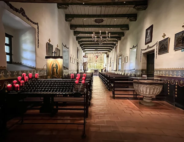 Interior view of Serra Chapel at Mission San Juan Capistrano, showing pews, red prayer candles, and a gold altar at the far end.