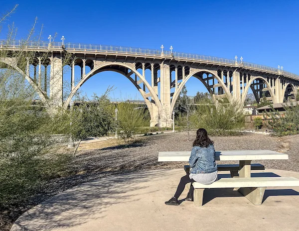 woman sitting at picnic table in a park admiring a bridge