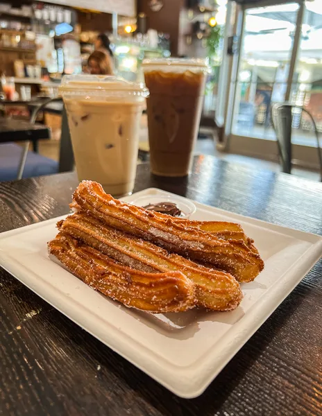 late of Spanish-style churros with a cup of chocolate dipping sauce, accompanied by two iced coffee drinks in the background at Amara Café in Pasadena.