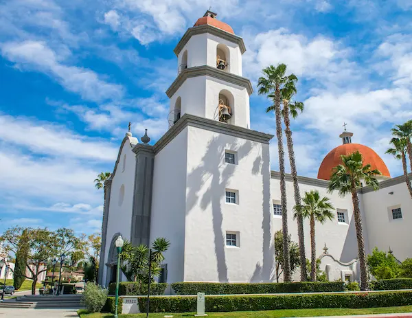 Exterior view of Mission Basilica San Juan Capistrano on a sunny day, with tall palm trees and a red dome contrasting against the white building.