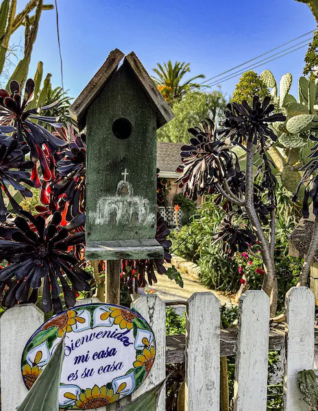 A green birdhouse and colorful ceramic “Bienvenida, mi casa es su casa” sign on a white picket fence, surrounded by succulents and cacti.