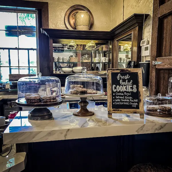 A marble countertop at Trevor on the Tracks showcasing fresh-baked cookies under glass domes, with a chalkboard sign listing cookie flavors.