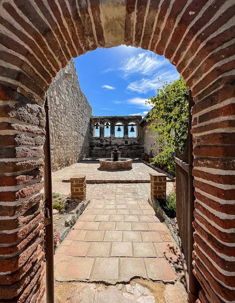 A brick archway leading into the Sacred Garden at Mission San Juan Capistrano, with a view of the historic bell wall and crosses.