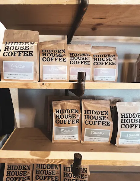 Bags of Hidden House Coffee displayed on wooden shelves, featuring different roasts like “Mexico” and “Ecuador.”