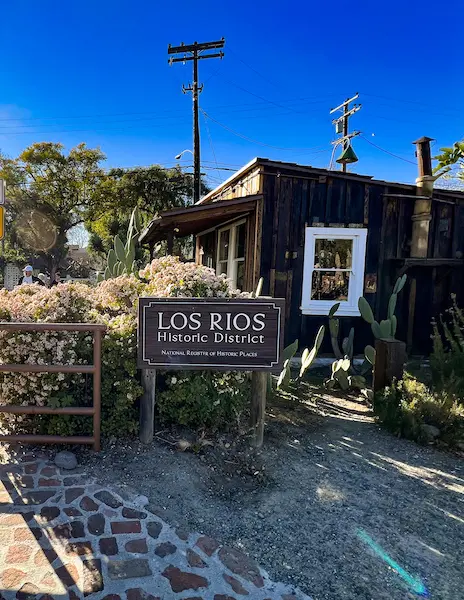 A wooden sign marking the entrance to the Los Rios Historic District, with blooming flowers, cactus plants, and a rustic wooden building in the background.