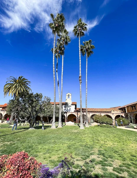 View of the main courtyard at Mission San Juan Capistrano, with tall palm trees, green lawn, and the surrounding mission buildings.