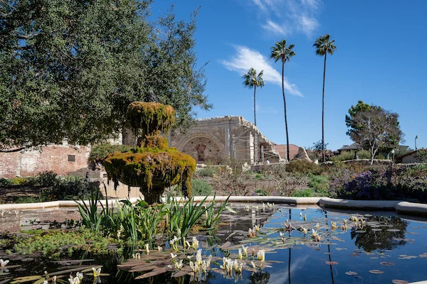 A moss-covered fountain in the garden at Mission San Juan Capistrano, with lily pads in the water and ruins of the Great Stone Church in the background.