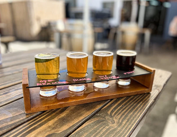 Flight of four craft beers on a wooden tray, labeled with chalk, sitting on an outdoor picnic table at Overtown Brewery.