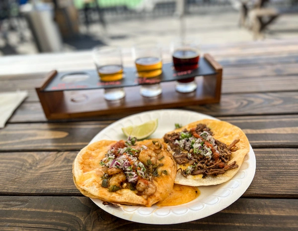 A plate with two street tacos served with a lime, with a flight of beer tastings in the background, set on a wooden table.