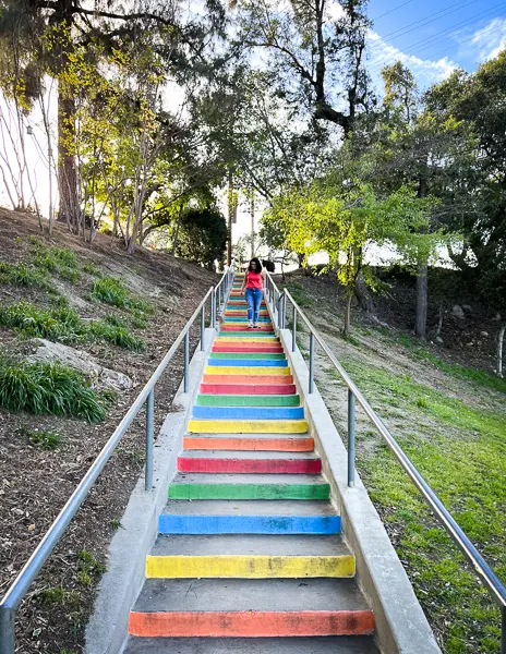 Person (kathy) walking up a colorful set of rainbow-painted stairs at Kiwanis Park, surrounded by trees and greenery.