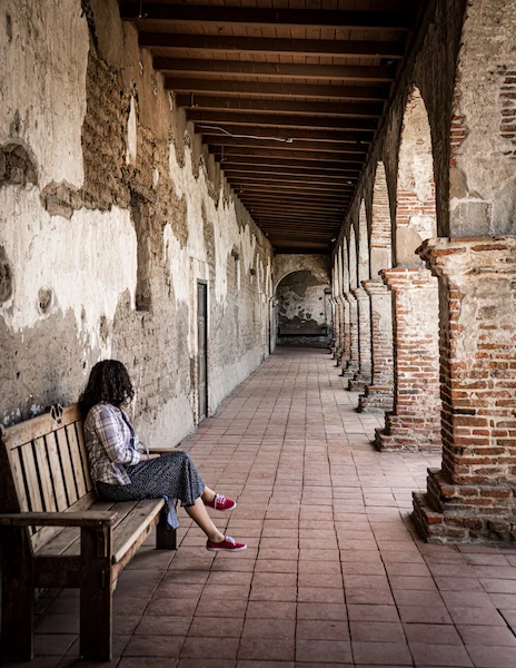 A woman (kathy) sitting on a bench along the corridor at Mission San Juan Capistrano, with weathered adobe walls and arched columns.
