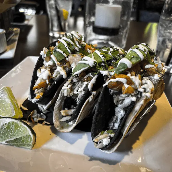 Three grilled fish tacos topped with avocado, shredded cheese, and crema on a white plate with lime wedges, served at Trevor on the Tracks.