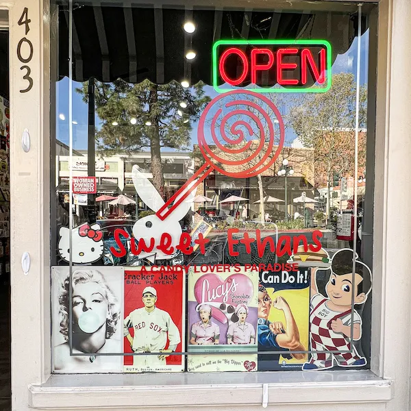Sweet Ethans storefront window with neon "OPEN" sign, retro candy signs, and classic pop culture graphics.