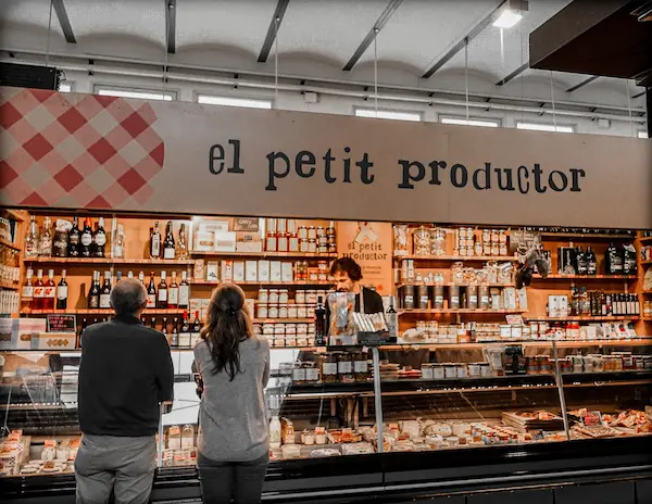 El Petit Productor stall at Mercat del Lleó in Girona, Spain, showcasing local cheeses, wines, and artisanal goods with customers browsing