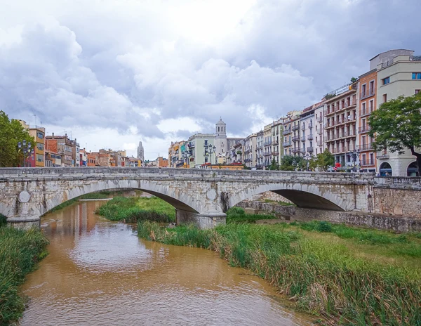 Stone bridge over the Onyar River in Girona, Spain, with colorful buildings and the Girona Cathedral in the background under a cloudy sky.