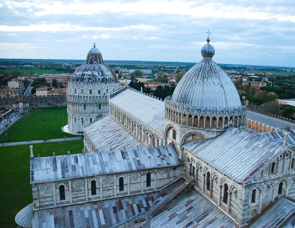 view of the Pisa Cathedral and the Baptistery in the Piazza dei Miracoli. The intricate marble facades and domed roofs of these historic buildings are highlighted, set against the backdrop of Pisa's expansive landscape. The green lawns of the square add a vivid contrast to the grey tones of the structures.