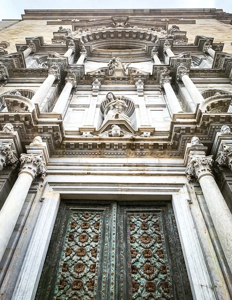 A close-up view of the ornate entrance to Girona Cathedral, highlighting its intricate Baroque facade, towering columns, and detailed bronze doors.